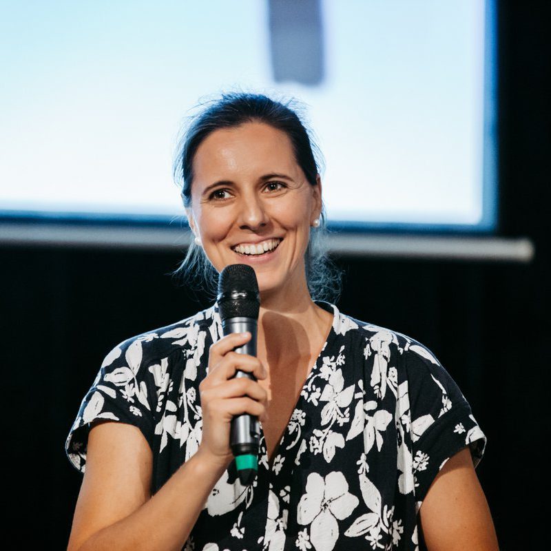 Lea Boettger, lächelnd mit Mikrofon. Frau mit floralem Oberteil.