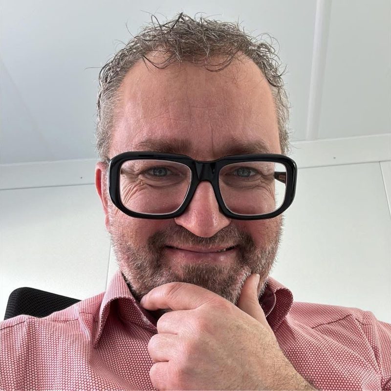 Porträt von Markus Leimegger mit Brille, lächelnd, die Hand am Kinn. Freundlicher Gesichtsausdruck.