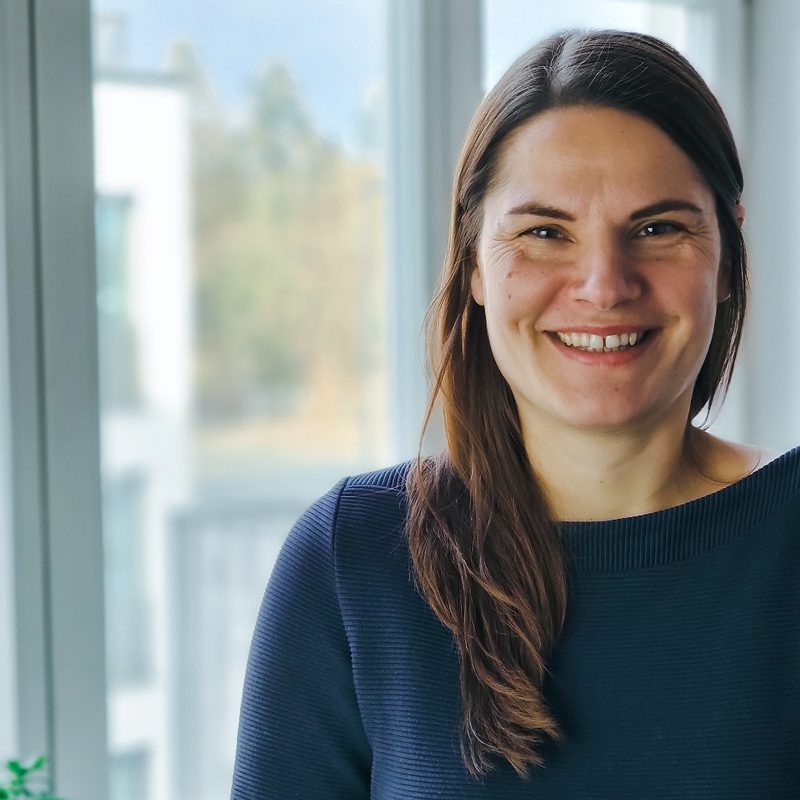 Kathrin Hausberg Portrait: Frau lächelt in die Kamera, dunkles, langes Haar, blauer Pullover.