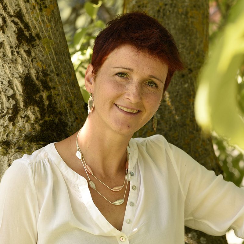 Foto Ruth Rieder: Frau mit kurzem, rotem Haar, weißer Bluse und Halskette lächelt vor Baum.