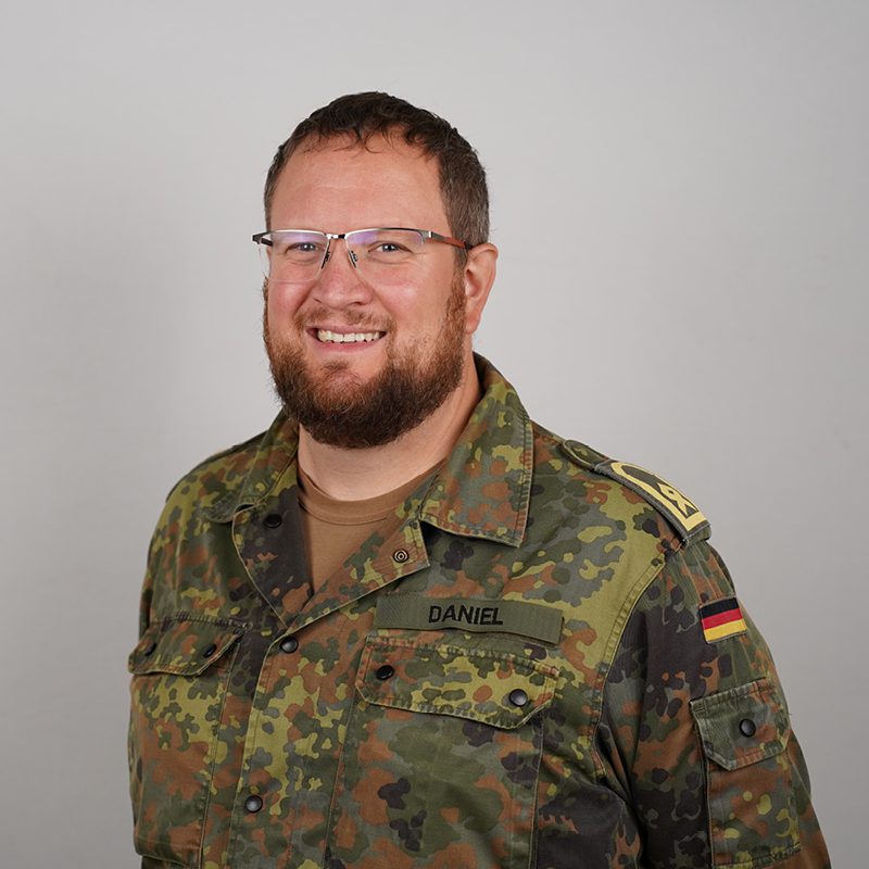 Porträt: Soldat Daniel in Uniform, mit Brille und Bart, lächelt. Foto 800x800px.