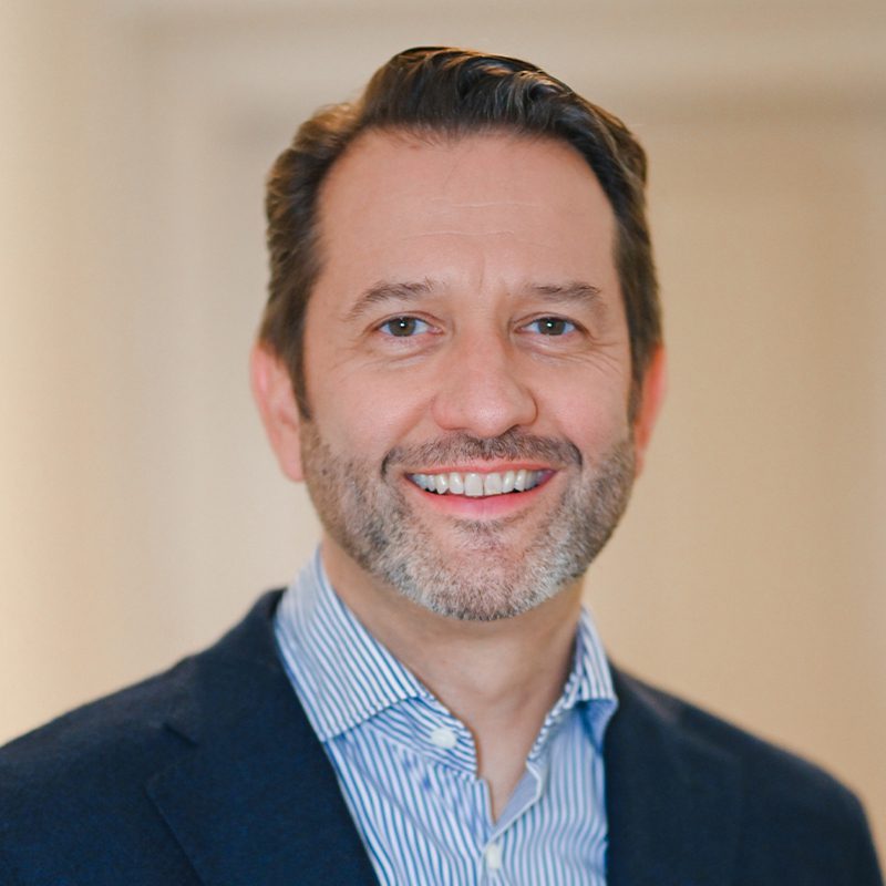 Porträt von Johannes Ermatinger, lächelnd, mit Bart und blau gestreiftem Hemd unter einem dunklen Jackett. Freundliches Aussehen.